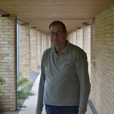 unser Schriftführer steht in einem langen, modernen Korridor mit gelben Ziegelwänden. Er trägt ein graues Polohemd und sieht freundlich in die Kamera. Um ihn herum sind Pflanzen und eine klare Gehfläche. Der Korridor vermittelt ein lichtdurchflutetes und offenes Gefühl.