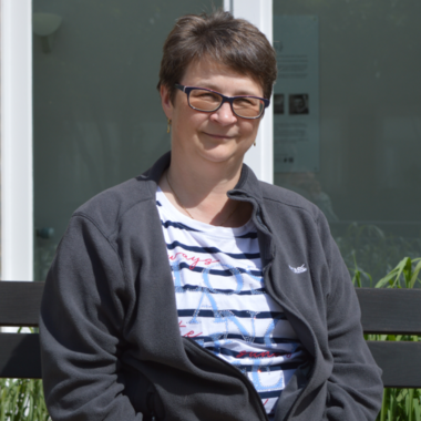 unsere Webmasterin sitzt auf einer Bank und lächelt. Sie trägt eine graue Jacke und ein gestreiftes Shirt. Im Hintergrund sind grüne Pflanzen und ein helles Fenster zu sehen.