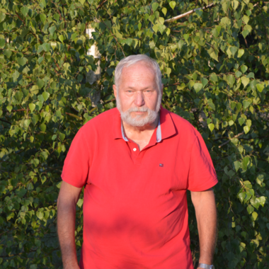 unser Beisitzer steht vor einem Hintergrund aus grünen Blättern. Er trägt ein rotes T-Shirt und schaut direkt in die Kamera. Die Atmosphäre wirkt freundlich und entspannt.